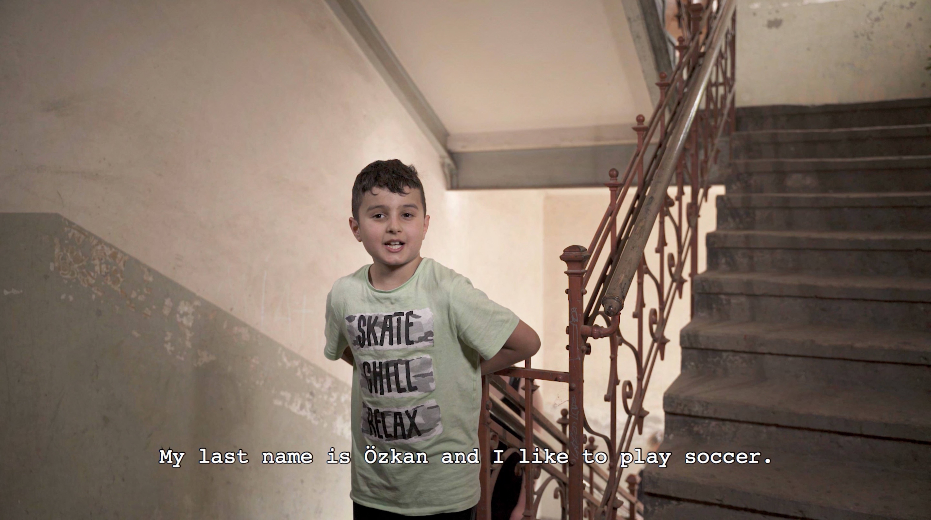 A young boy stands in an apartment block stairwell. A subtitle reads "my last name is Ôzkan and I like to play soccer"