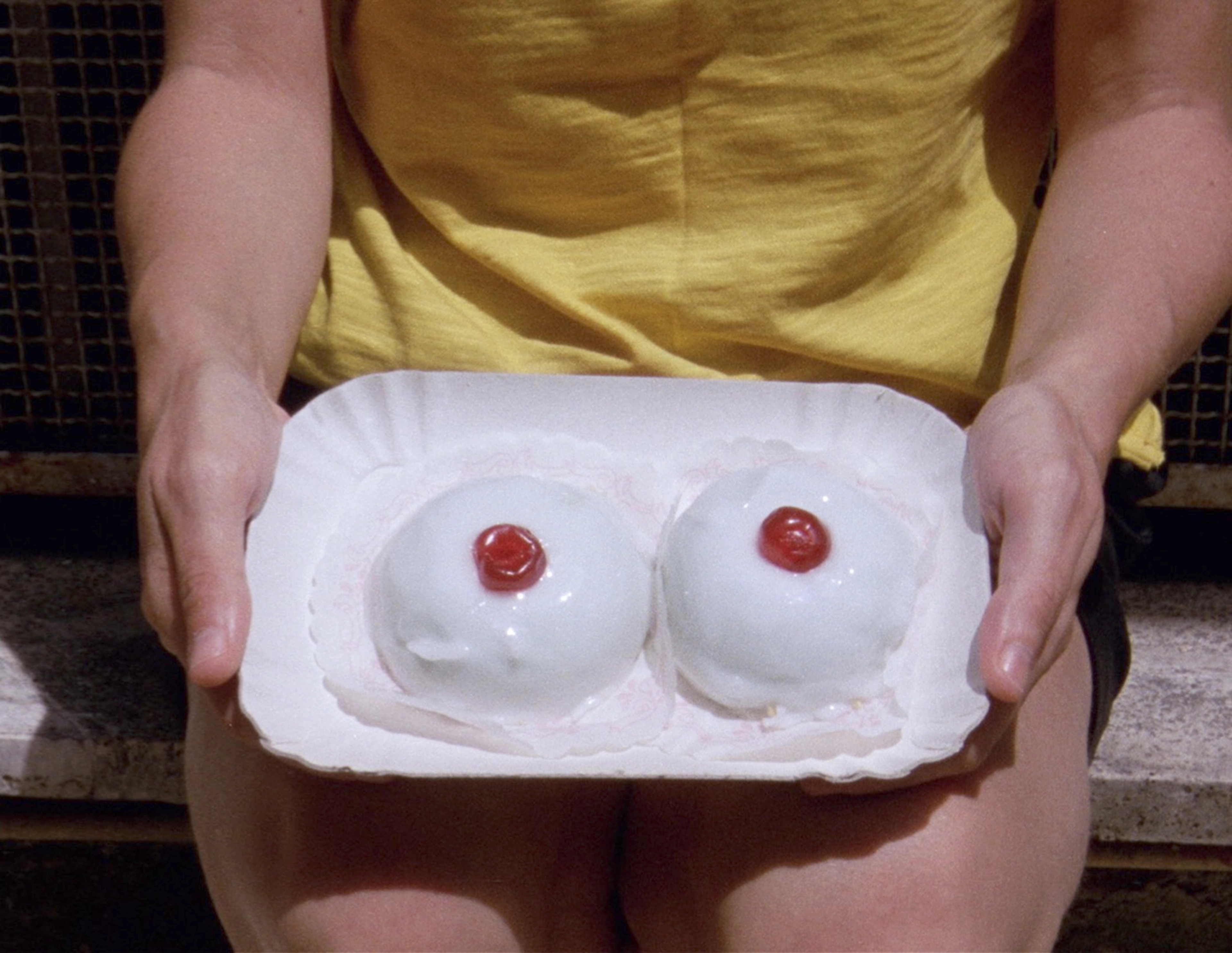 A woman in yellow sits holding a rectangular plate with two white circular desserts with cherry nipples.