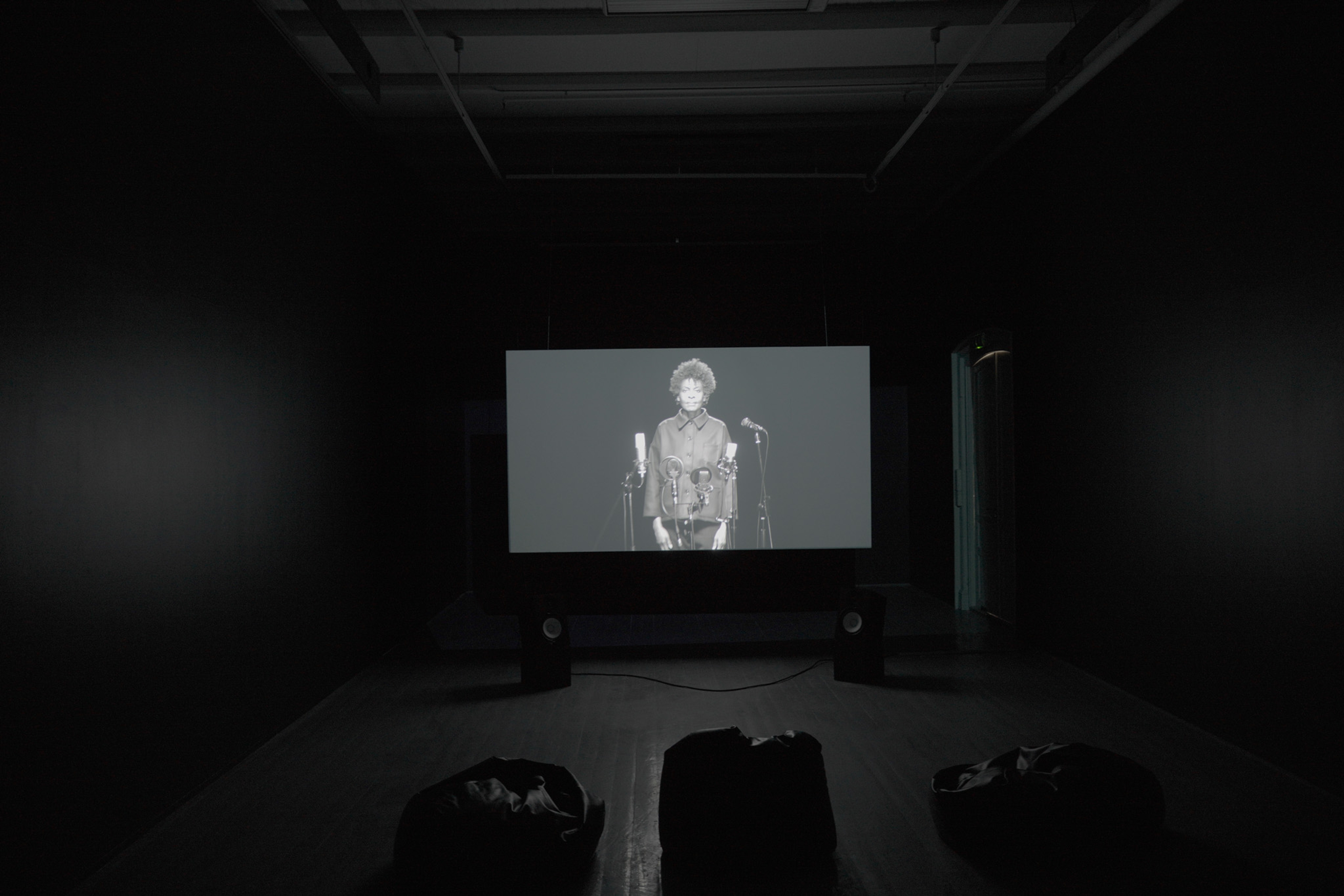 En projeksjonslerret vises en kvinne som står foran mikrofoner i svart-hvitt.