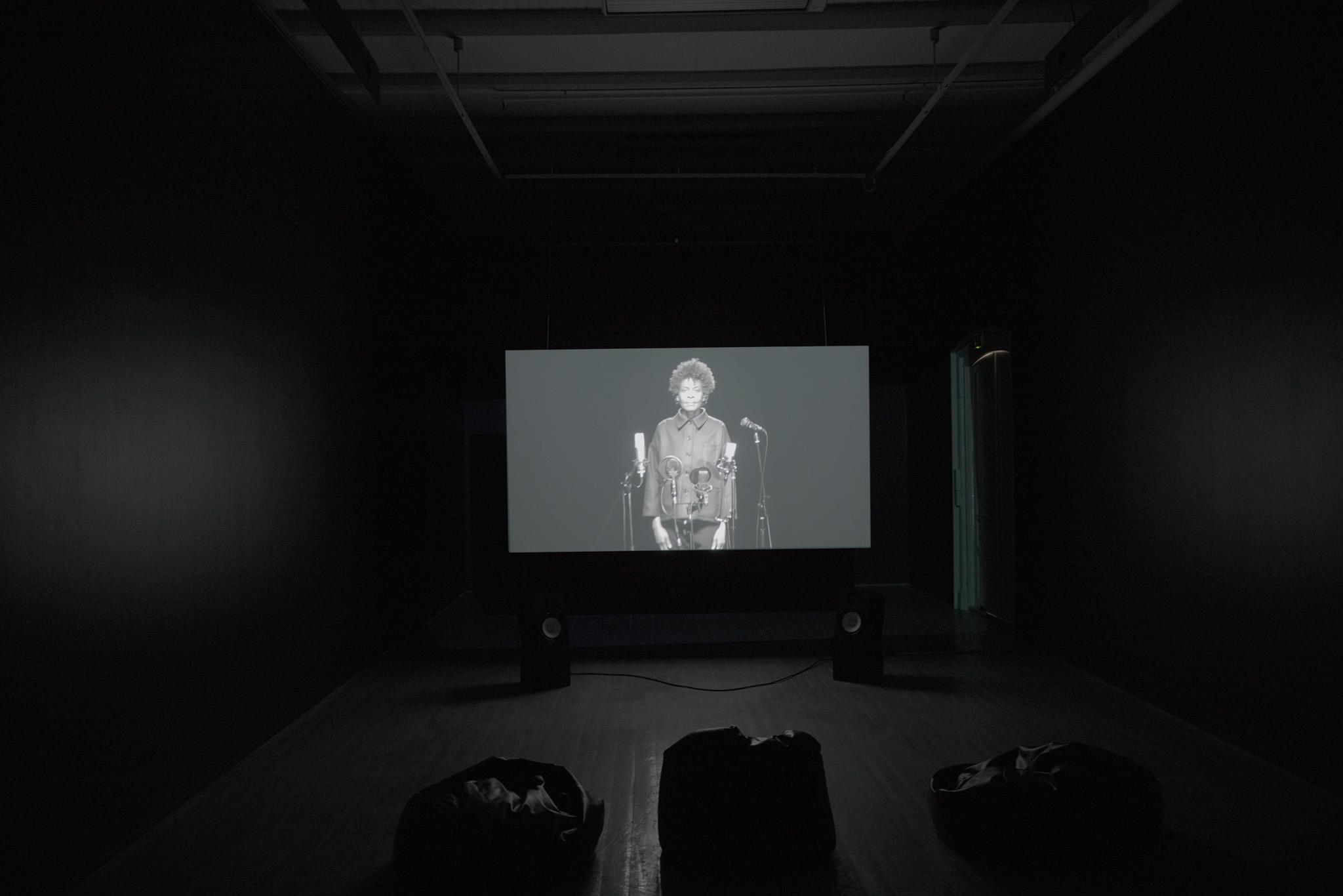 En projeksjonslerret vises en kvinne som står foran mikrofoner i svart-hvitt.