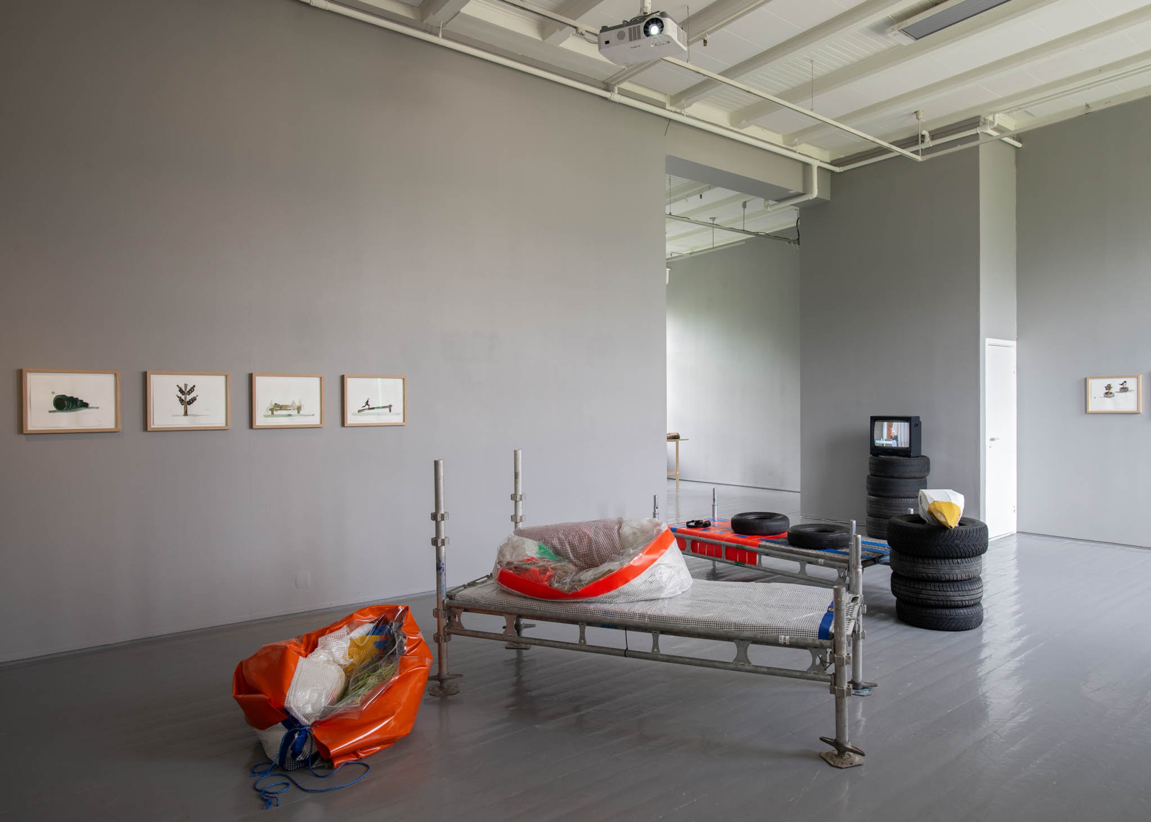 A scaffold bench stands in the middle of a gallery. Drawings hang on the wall, and there is an old tv in the background.