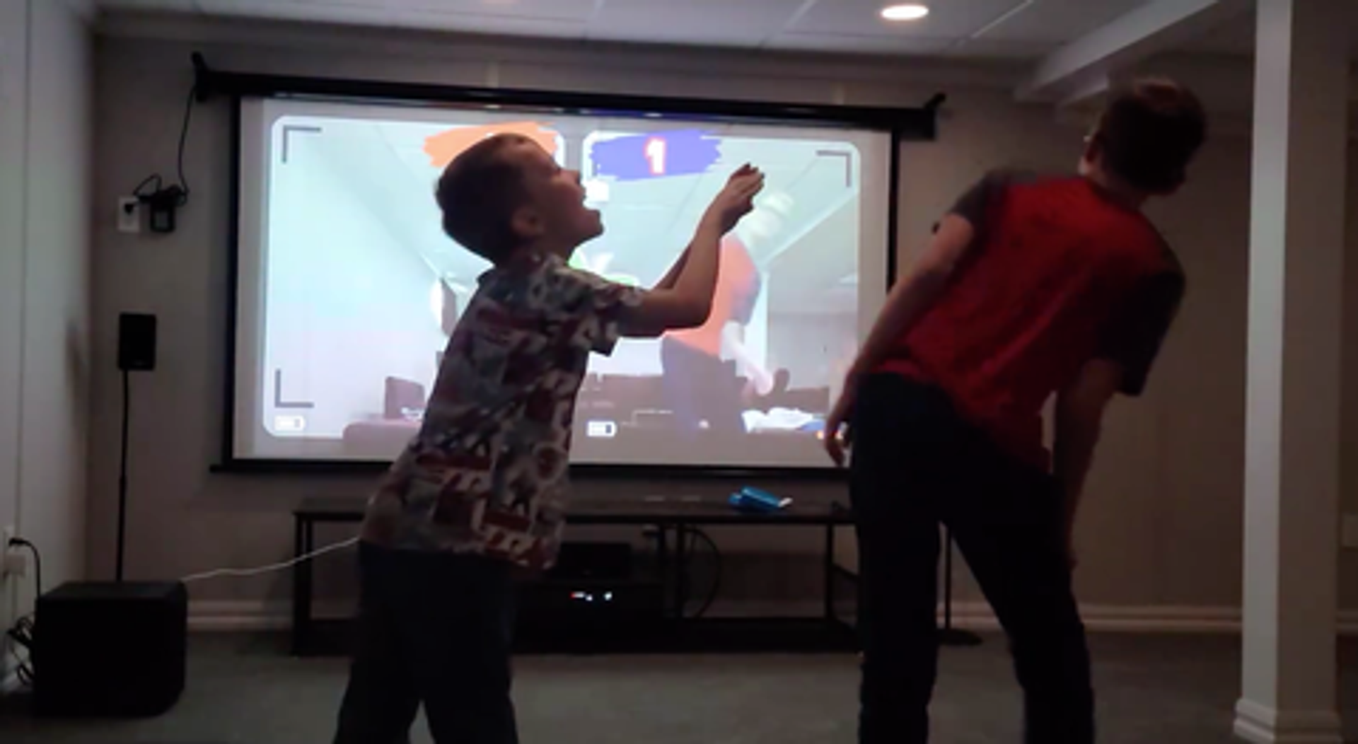 A younger boy laughs and reaches his hands up while playing a split-screen active video game next to his older brother on a projector connected to the Nex Playground device.