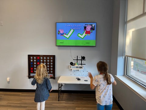 Two young girls playing a motion-controlled Peppa Pig active game on the Nex Playground console to ease anxiety while waiting at the Eric's RX clinic.