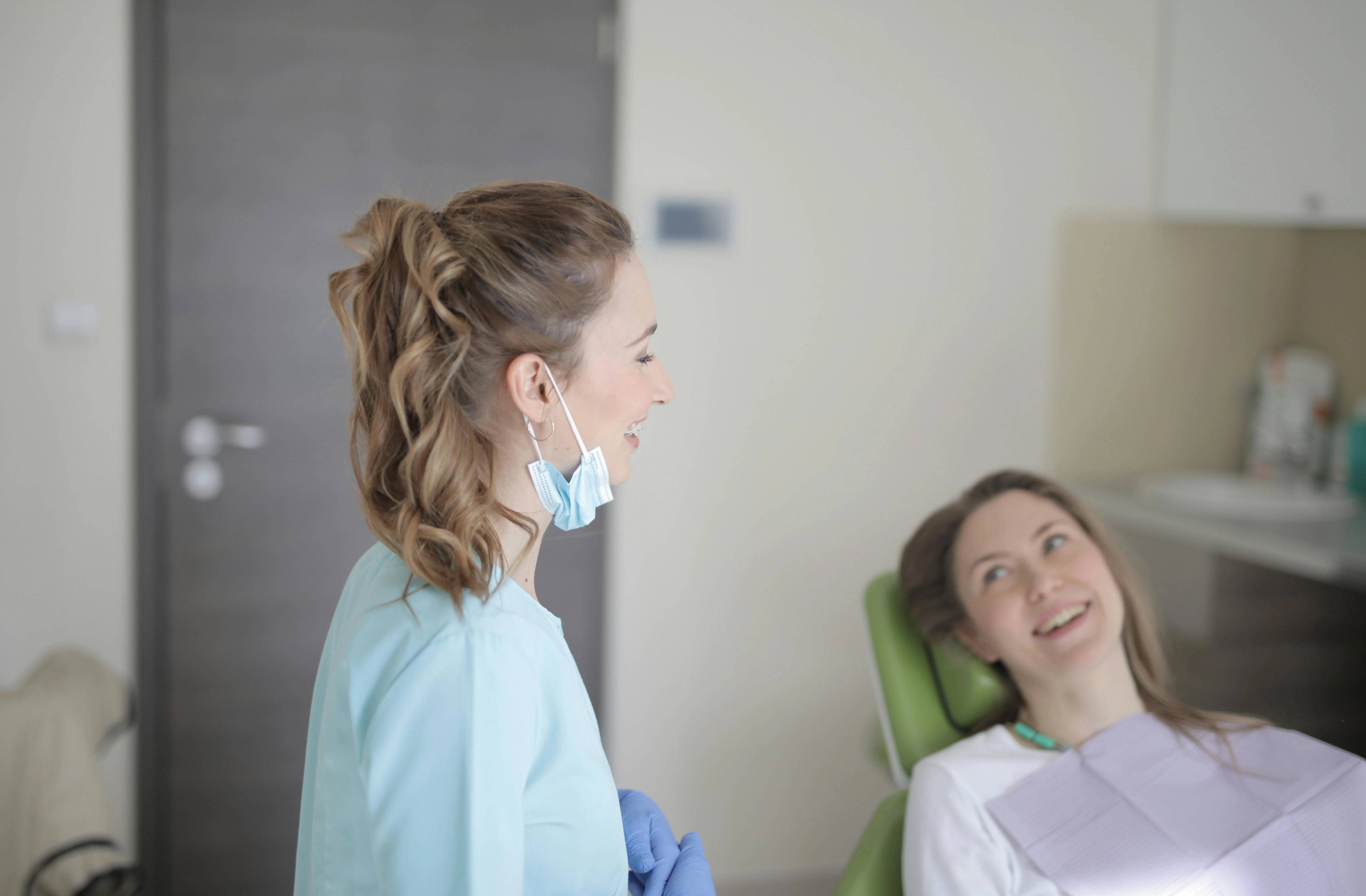 A stock photo of a patient smiling towards her dentist who is wearing a face mask pulled down below her chin