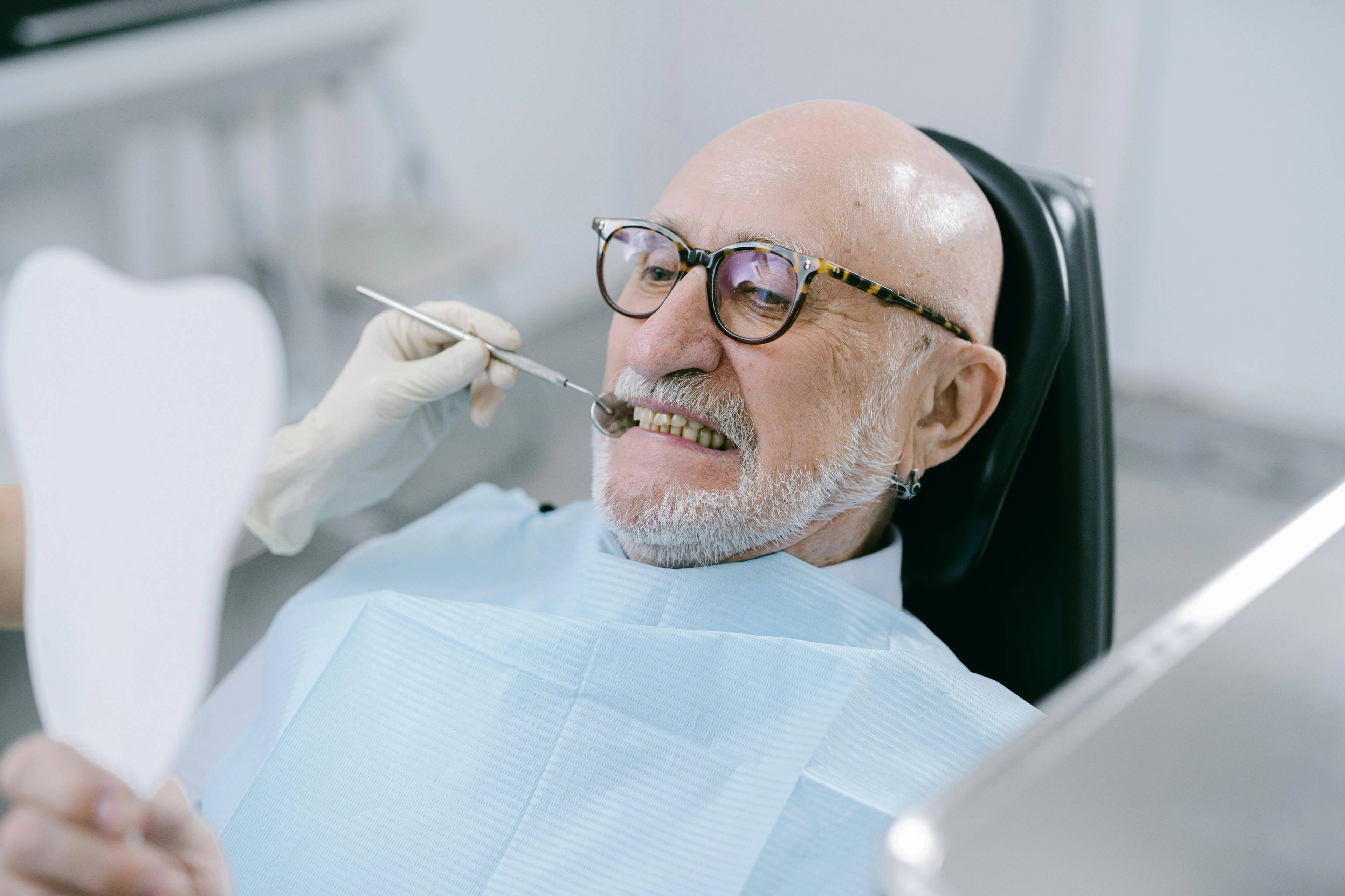 An older male patient wearing glasses has his teeth examined by a dentist wearing medical gloves