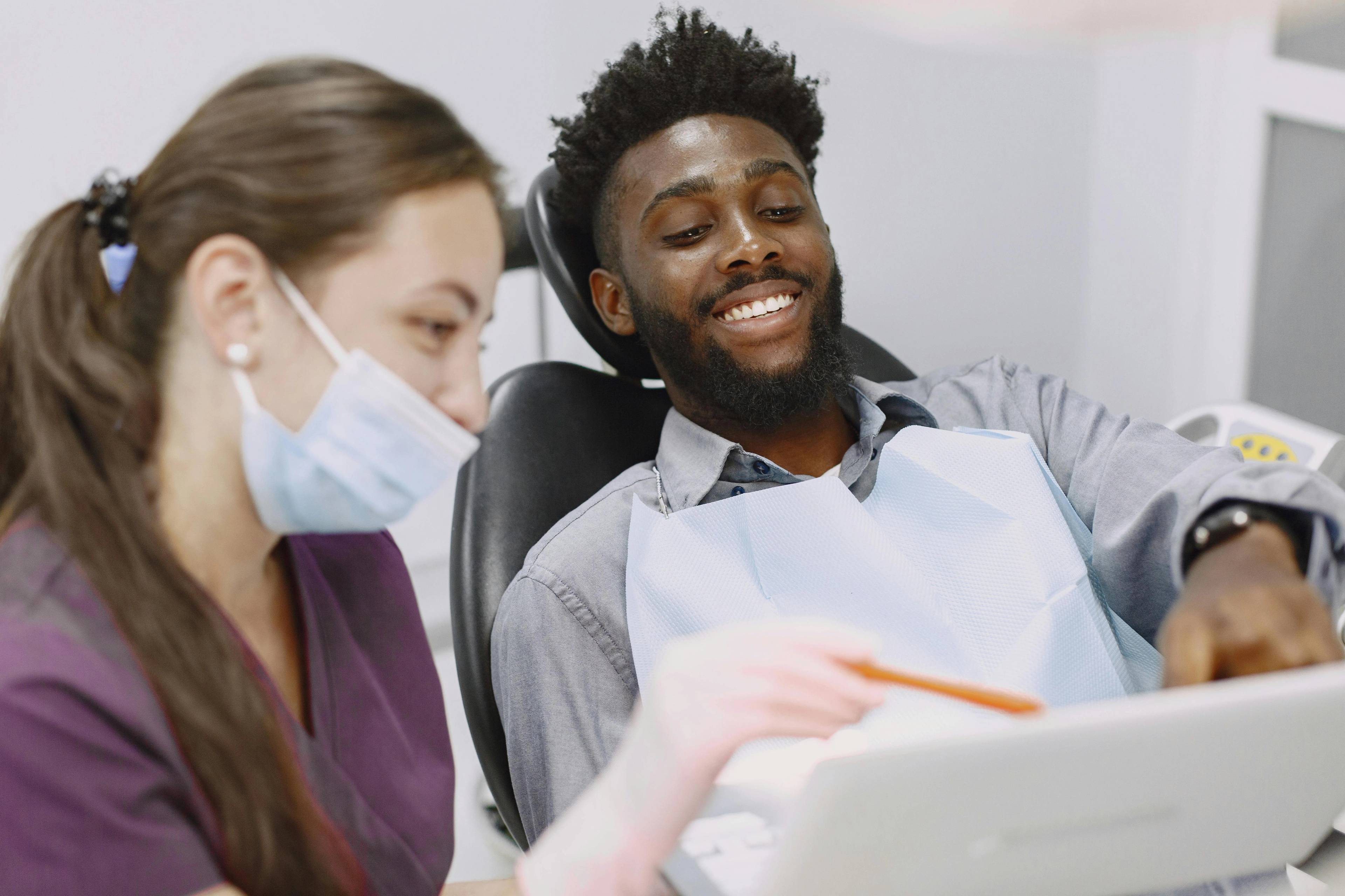 A stock photo of a male patient sitting in a dental treatment chair smiling with his dentist who is pointing to something on a screen that's out of frame