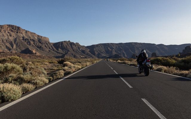 Motorcyclist riding on a long road through a desert landscape.