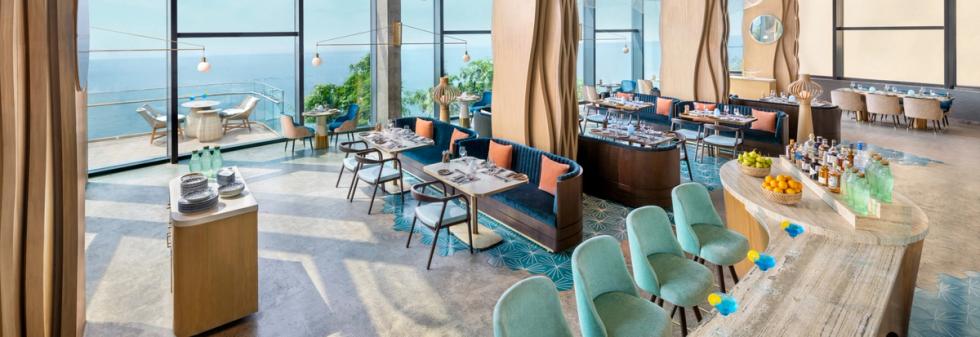 Restaurant with ocean-facing windows, curved wooden bar, and turquoise seating