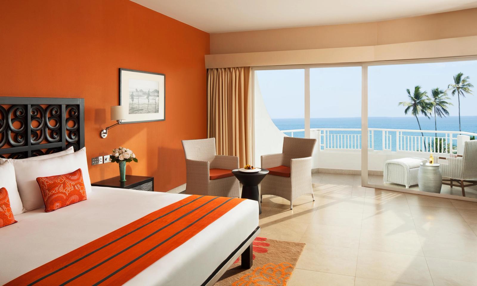 A room with an orange wall, a king bed and 2 armchairs by a glass door onto a balcony over the sea.