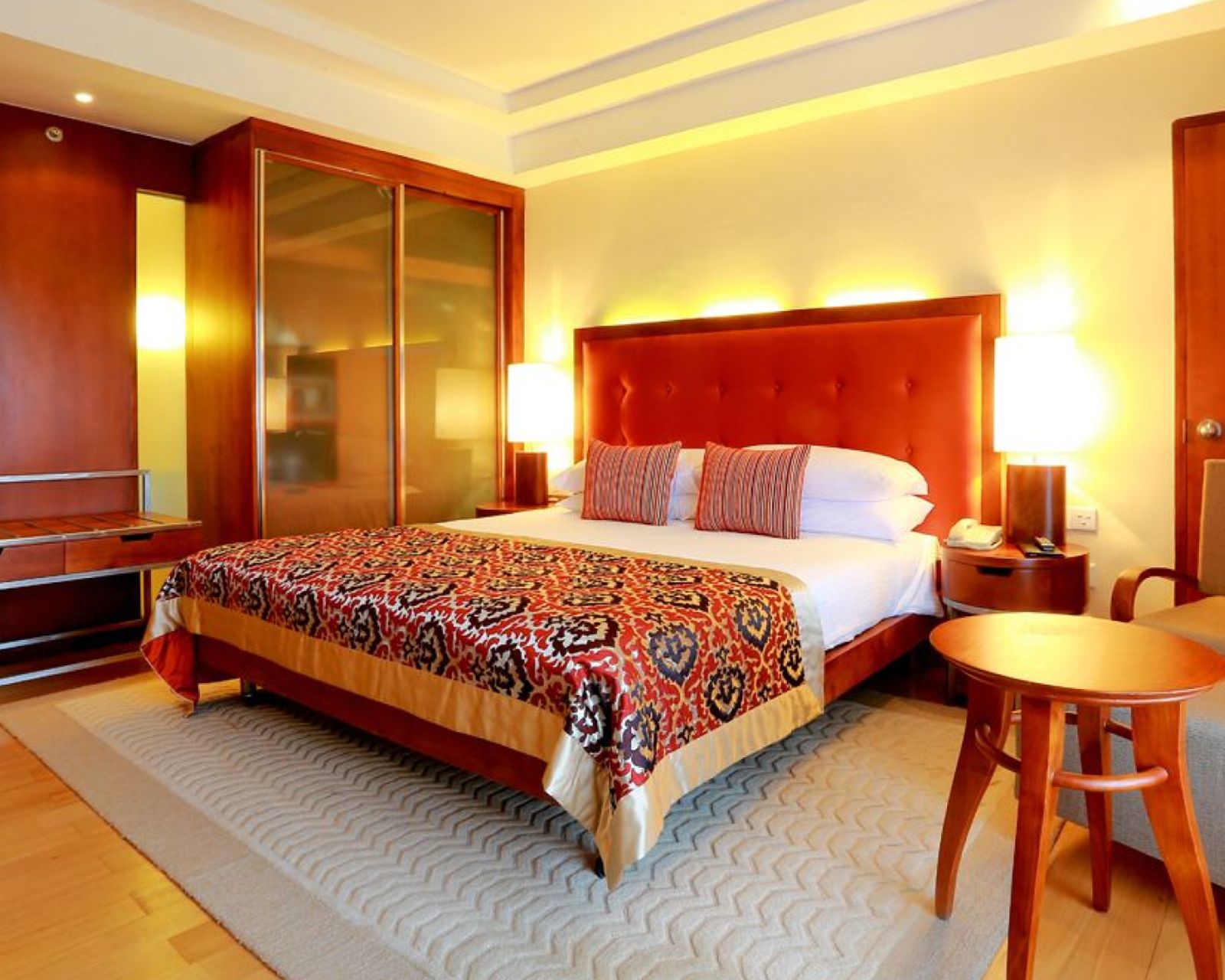 A Deluxe Room City Facing: a bed with a tall orange padded headboard and a red patterned throw, a wardrobe with glass doors in wood panelling, and a round wooden side table