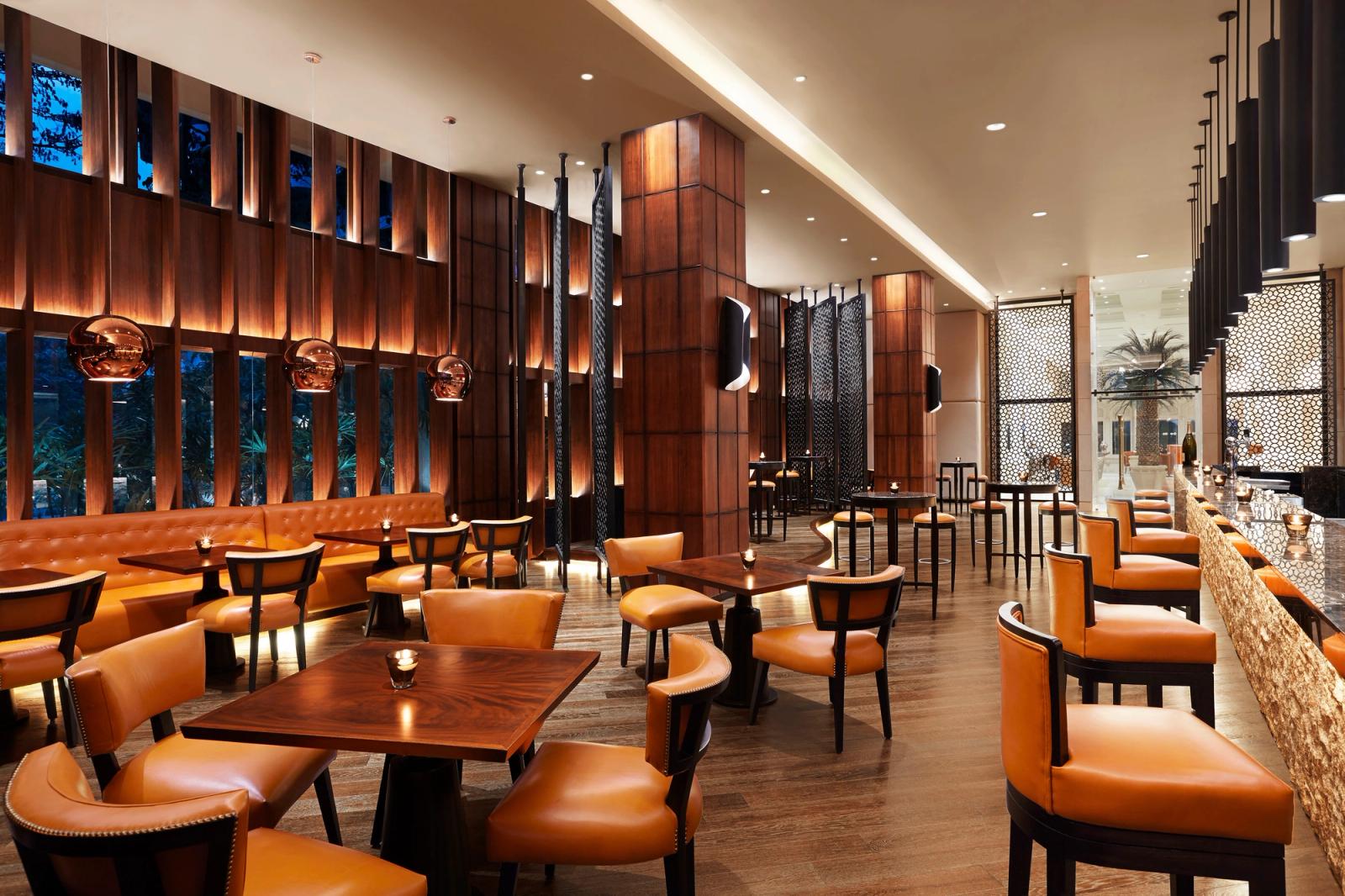 A restaurant and bar with orange leather chairs and a long banquette, wooden tables with candles, copper pendant lamps, and tall windows onto trees