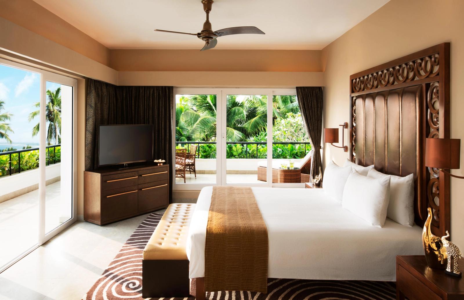 A suite bedroom with a carved headboard and glass doors onto a balcony above the palms.