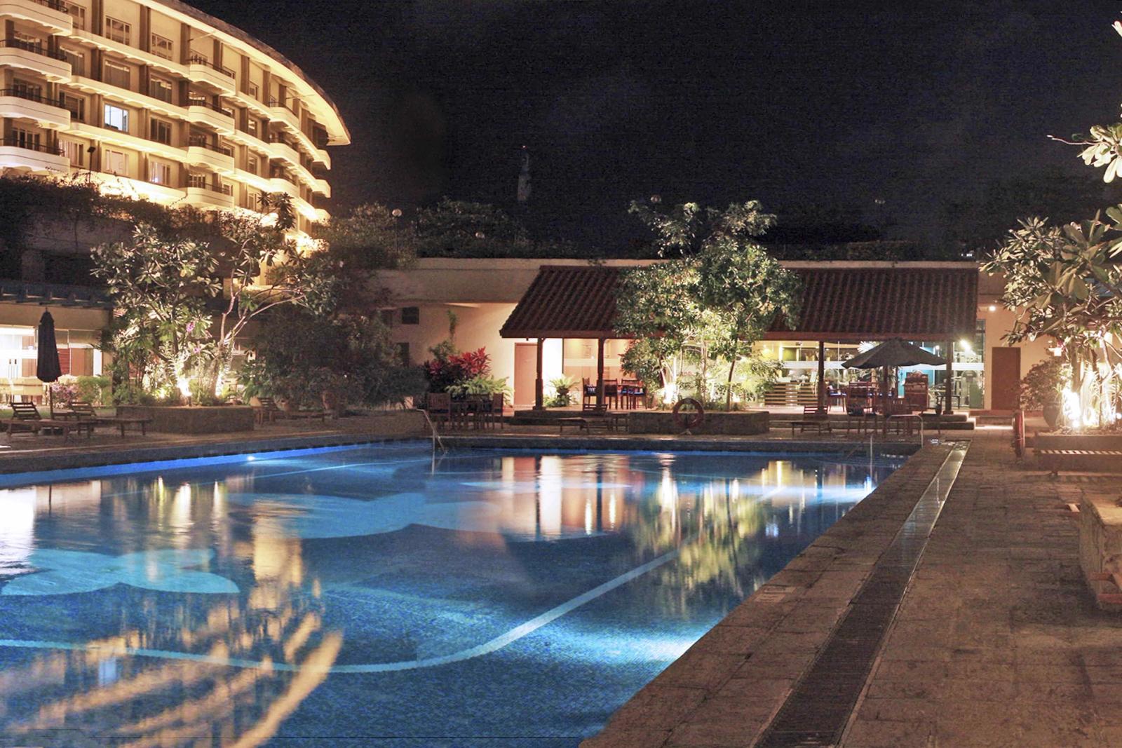 The outdoor pool at night, lit blue, with loungers along its edge, a pavilion under a tiled roof among frangipani trees, and a curved wing of rooms behind