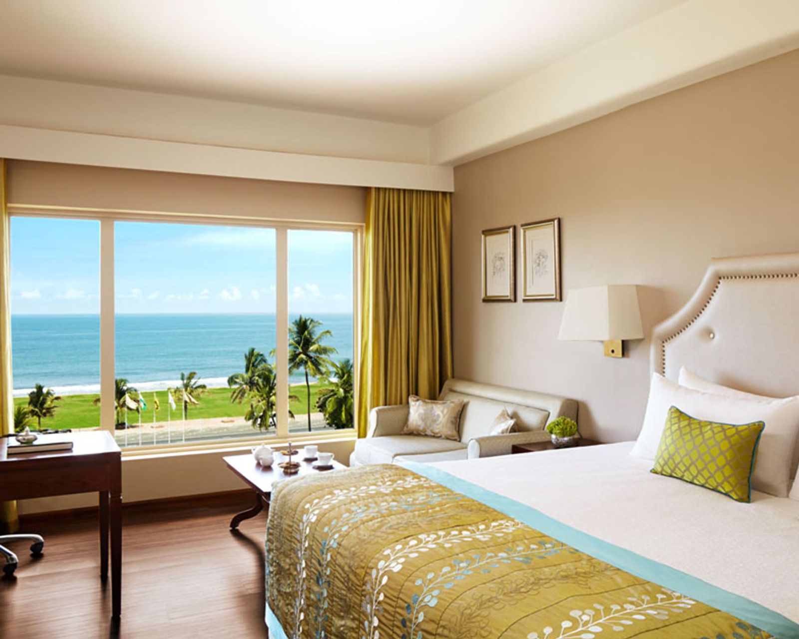 A Deluxe Room Ocean Facing: a bed with a padded headboard and a gold throw, a sofa and a tea table, and a wide window over a lawn with flagpoles, palms and the sea