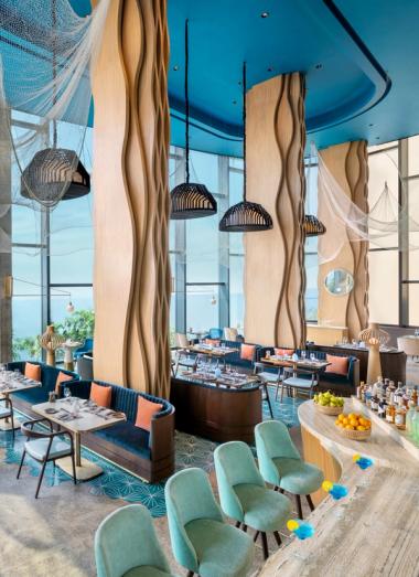 Restaurant with ocean-facing windows, curved wooden bar, and turquoise seating