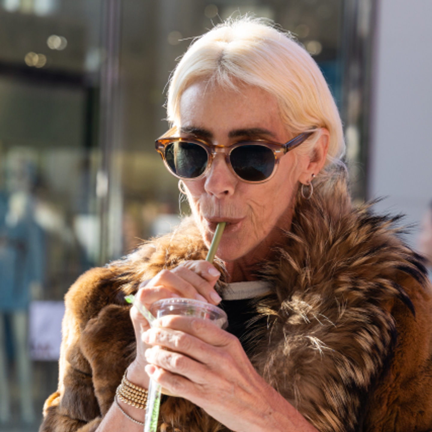 Woman in fur coat and sunglasses sipping drink through a straw.
