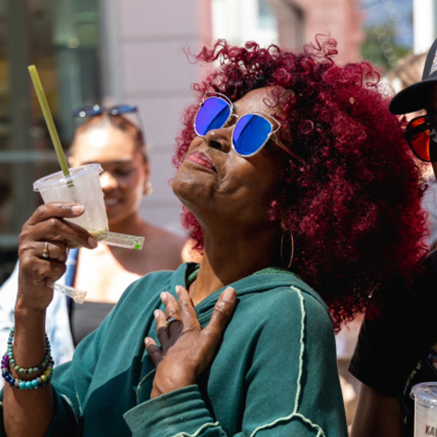 A woman with vibrant red hair, sunglasses, and bracelets enjoys a drink outdoors.