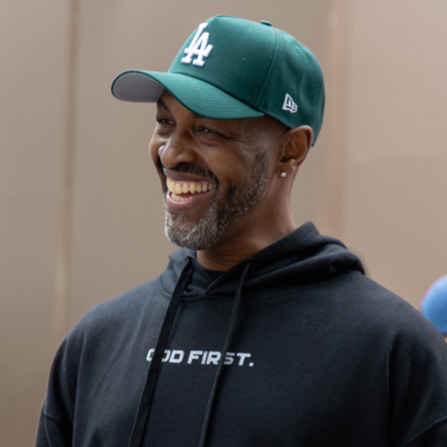 Man smiling, wearing a green cap and black hoodie with 'God First' text.