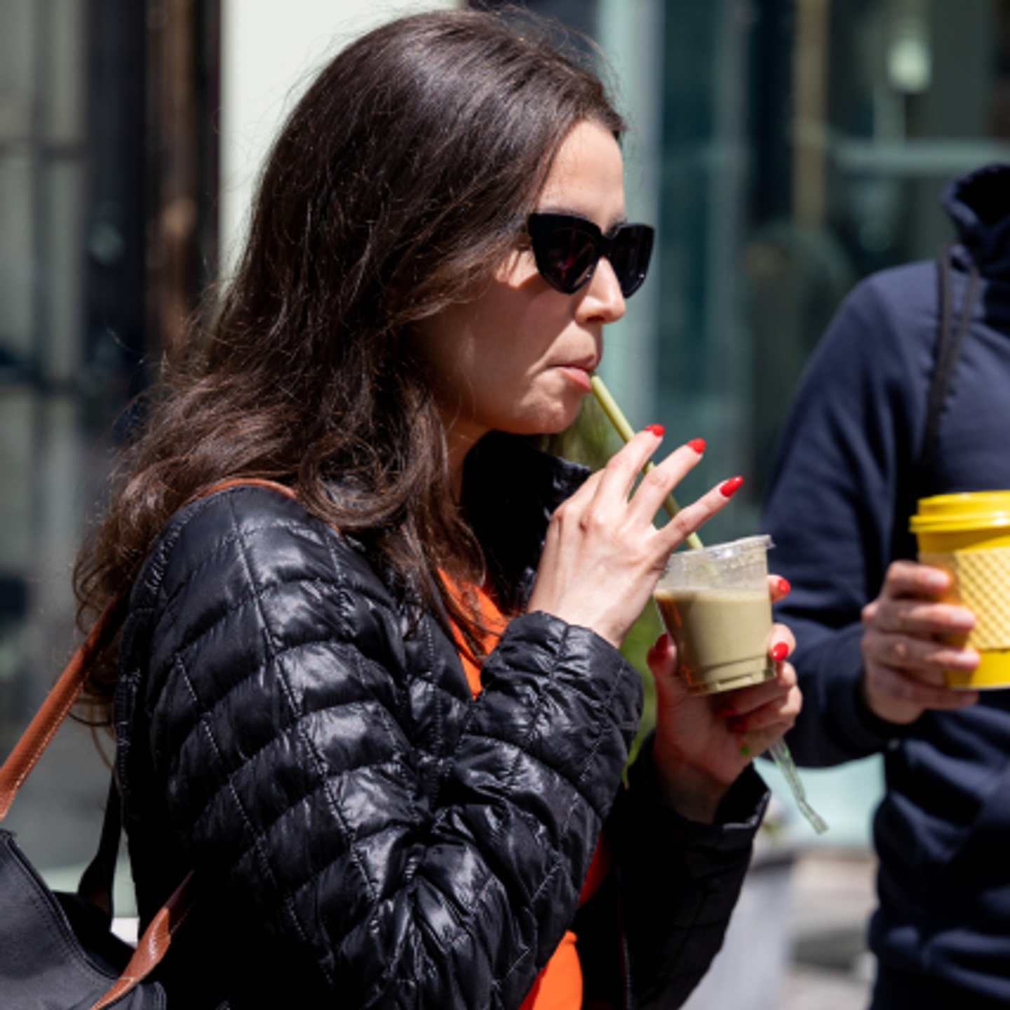 A woman drinks a smoothie while wearing sunglasses and a black jacket.