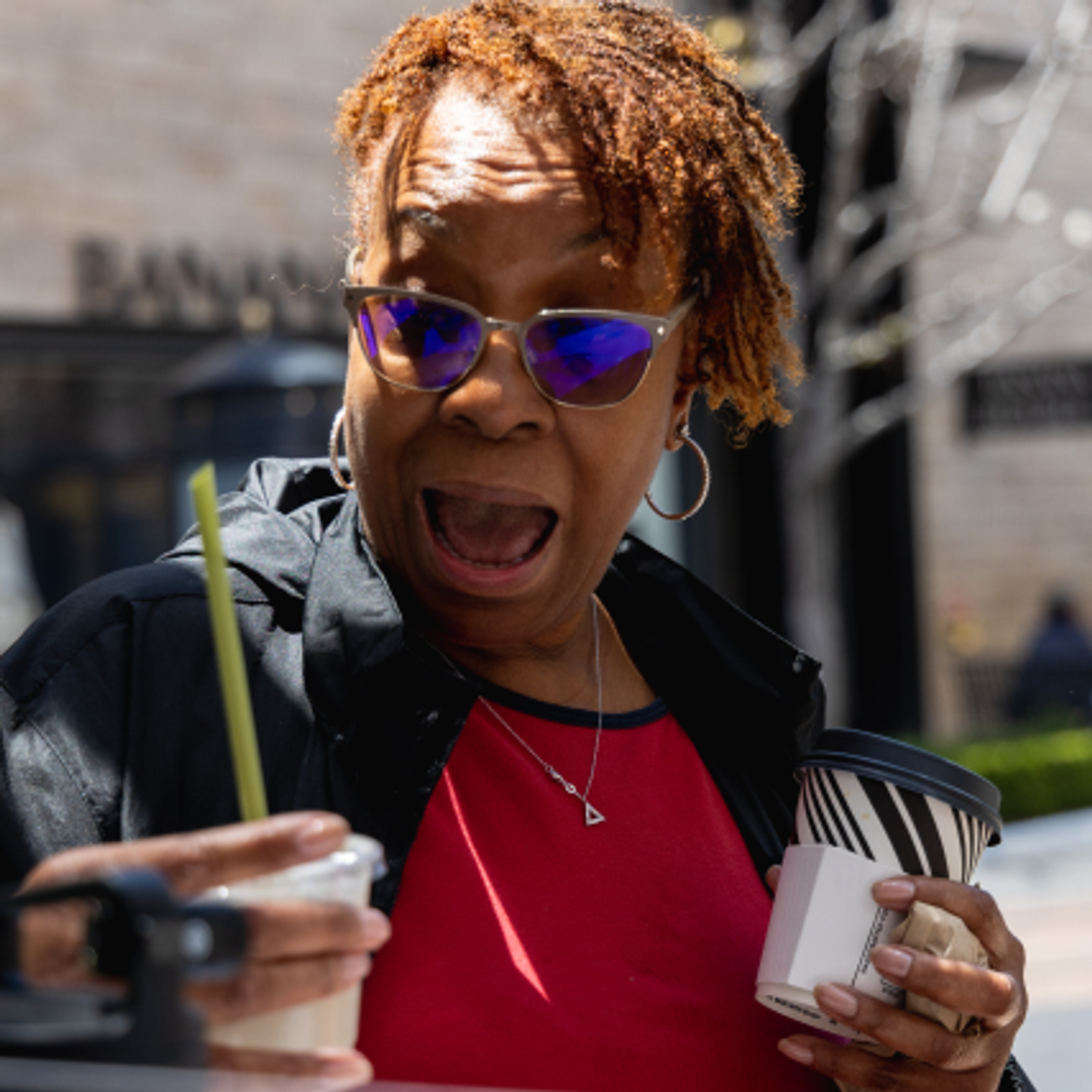 Surprised woman holding two beverages and wearing sunglasses outdoors.