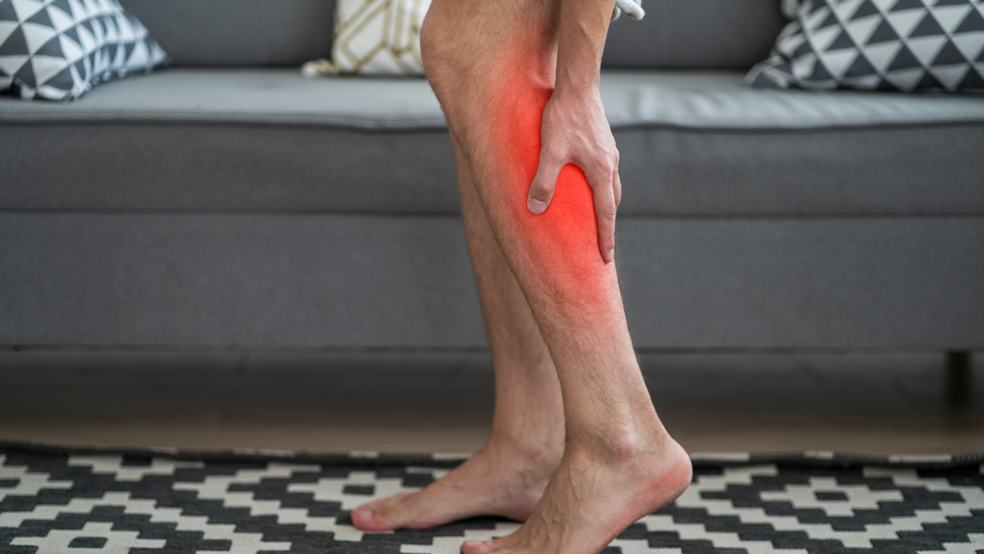 Person standing with one leg slightly bent, red glow on shin indicating leg pain or injury at home.