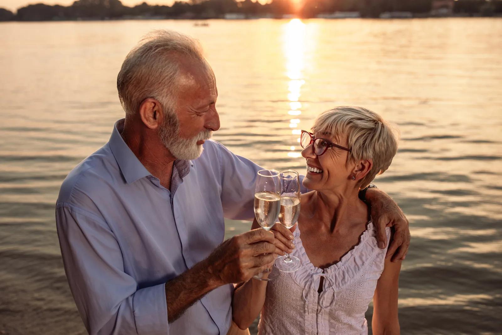 Un couple de seniors trinque au bord de l’eau au coucher du soleil.