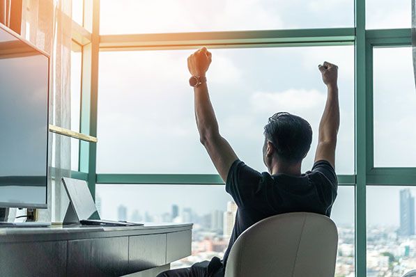 A person stretches both arms upward while seated at a desk facing tall windows.