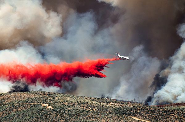 An aircraft drops red fire retardant over a smoke-covered hillside.