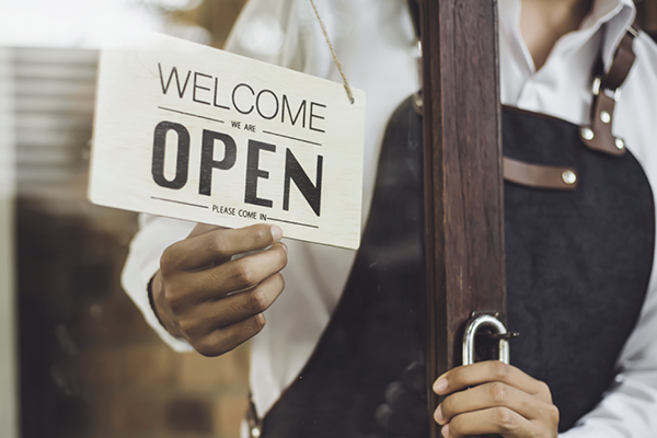 A shop worker holds a Welcome We Are Open sign and a padlock at a glass door.