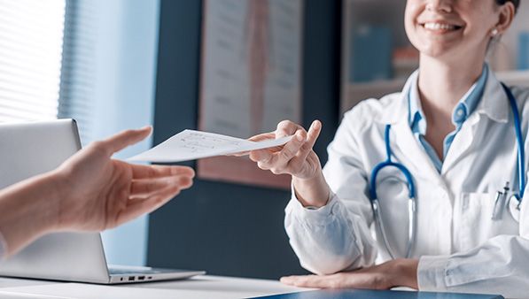 A clinician in a white coat hands a paper to someone across a desk.