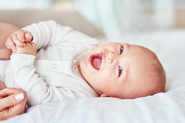 A baby in a white outfit lies on a bed with an adult's hand nearby.