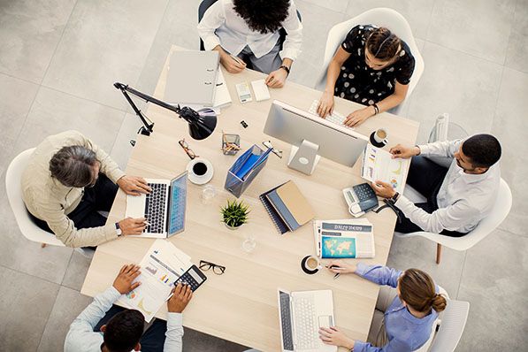 Coworkers work around a shared office table with computers and paperwork, viewed from above.