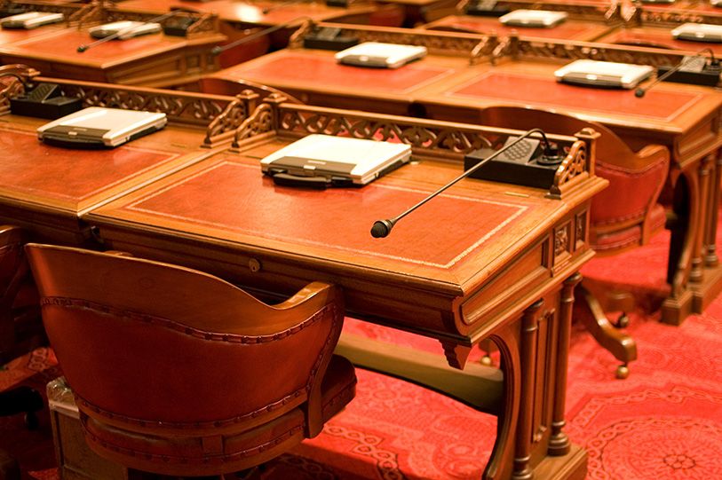 Rows of empty wooden desks and chairs on red carpeting in a legislative chamber.