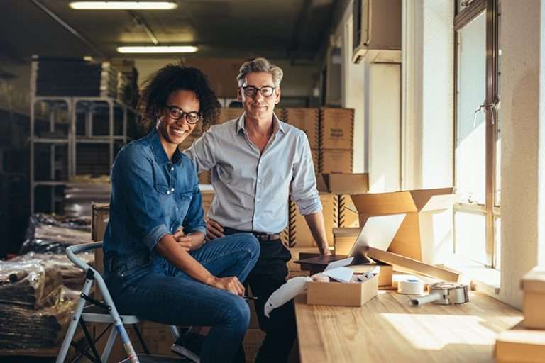 Two smiling people sit together at a workbench surrounded by boxes and workshop supplies.