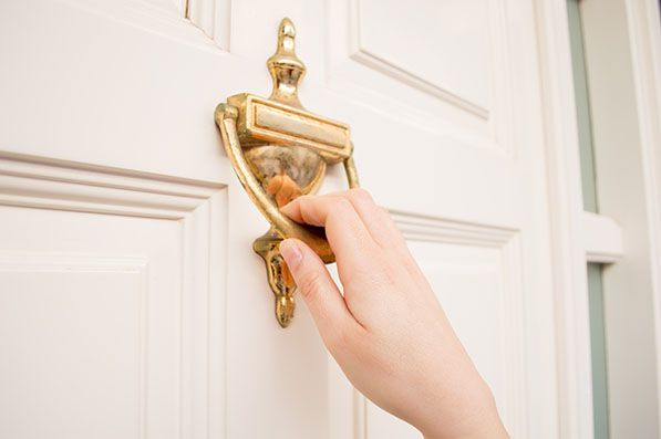 A hand holding a brass door knocker on a white door.