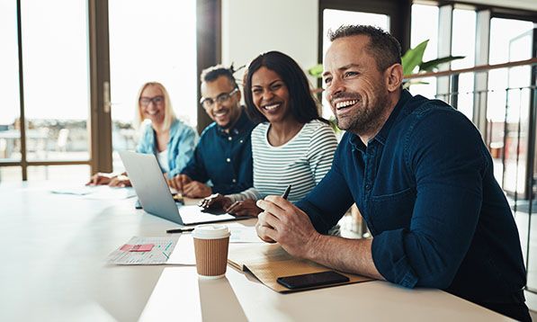 Four smiling coworkers sit at a long table with laptops and coffee cups.