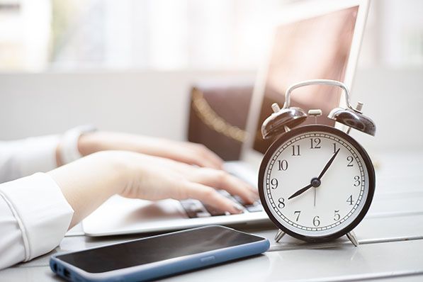 An alarm clock and smartphone sit beside someone typing on a laptop.
