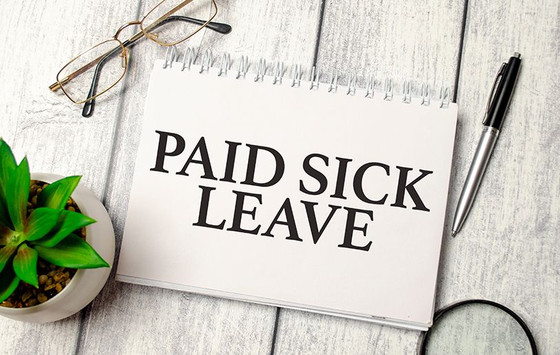 A notebook page reading Paid Sick Leave beside eyeglasses, a pen and a small plant.