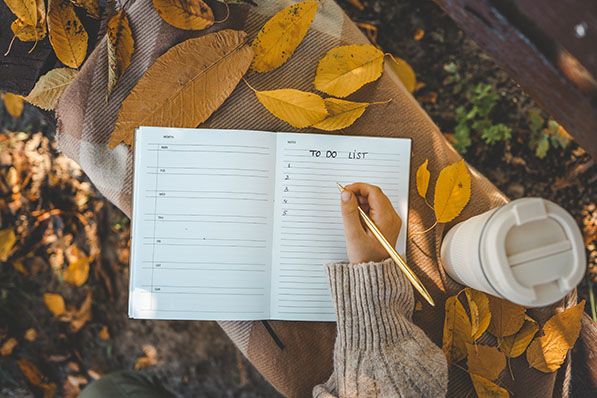 Hand writing a "To Do List" in a notebook on a plaid blanket, surrounded by autumn leaves and a travel mug.