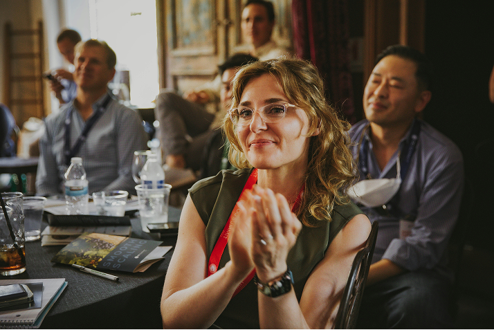 A smiling woman wearing glasses applauds while seated with other attendees at an event.