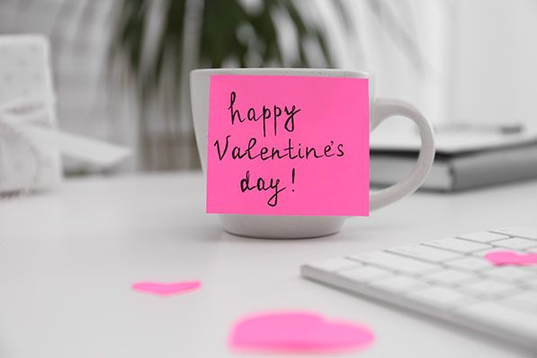 A mug bears a pink Happy Valentine's Day! note beside a keyboard and paper hearts.