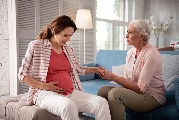 A pregnant woman holds her belly, grimacing slightly, while an older woman sitting beside her holds her hand with a concerned expression.