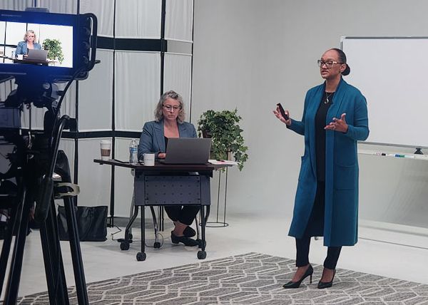 A woman presents in front of a video camera while another woman operates a laptop at a small table.