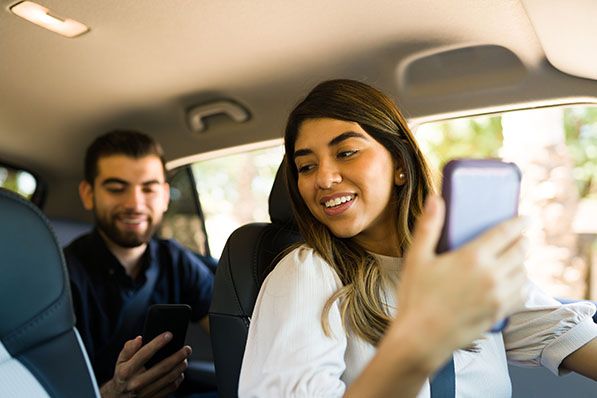 A woman in the front seat of a car looks at a phone while a passenger sits behind her.