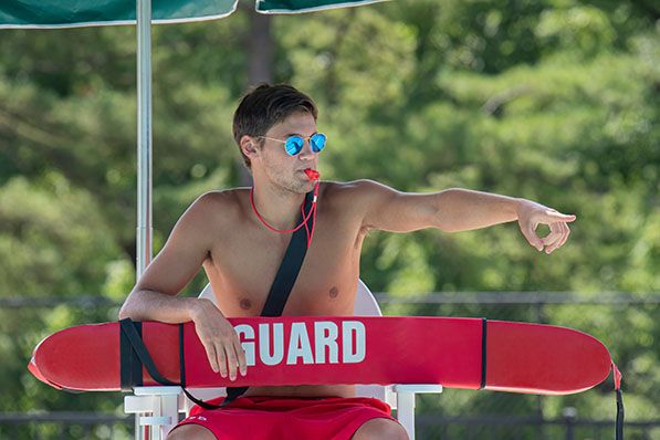 A lifeguard wearing sunglasses blows a whistle and points while holding a red rescue tube.