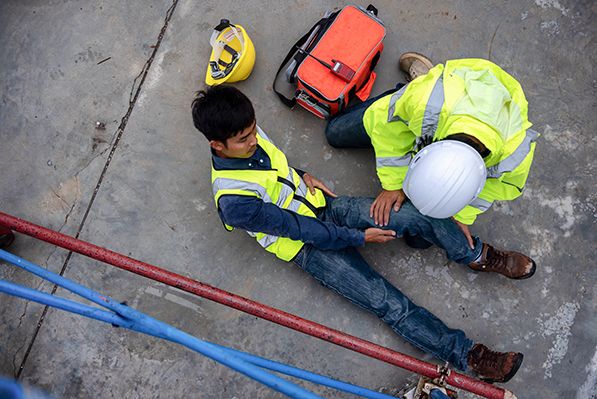 A worker in a hard hat kneels beside a seated coworker on a worksite floor near first-aid equipment.