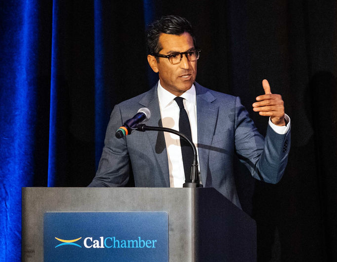 A man wearing glasses and a blue suit gestures while speaking at a CalChamber lectern.