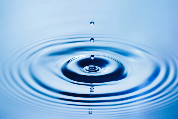 A water droplet falls into blue water, creating concentric ripples.