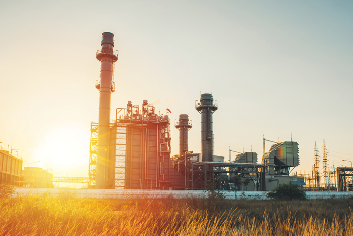 An industrial facility with tall smokestacks stands beside a grassy field at sunset.