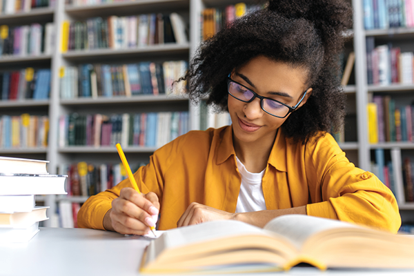 A student in glasses writes in a notebook beside an open book in a library.
